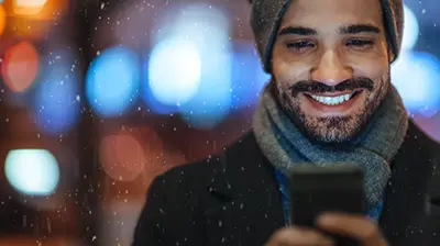 Have Yourself a Happy, Healthy Holiday Man dressed in winter clothes smiling as he looks at his phone while it snows