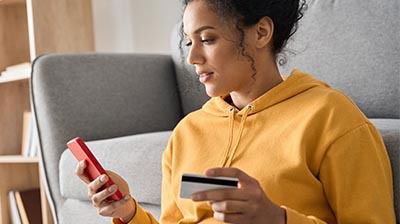 Your First Look – Applied Pay A woman in an orange hoodie using a red smart phone while holding a credit card with her other hand